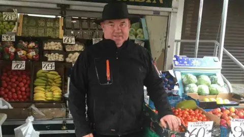 BEN COONEY Greengrocer Ben Cooney stands in front of his fruit stall. He is wearing a black fleece, black trousers and a black hat and smiling.