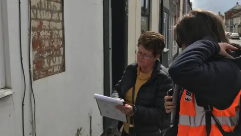MOLA Two volunteers standing on a pavement on a street of 19th Century-looking buildings, with the one in the middle holding a clipboard which she is looking at. She appears to be writing on the paper on the clipboard. The person to the far right is wearing a hi-vis jacket saying "MOLA".