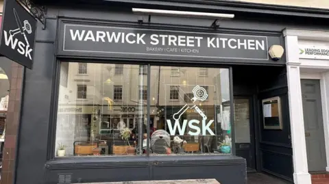The outside of a cafe called Warwick Street Kitchen. The shopfront is painted black with white lettering on the sign and a knife and mallet on the logo 