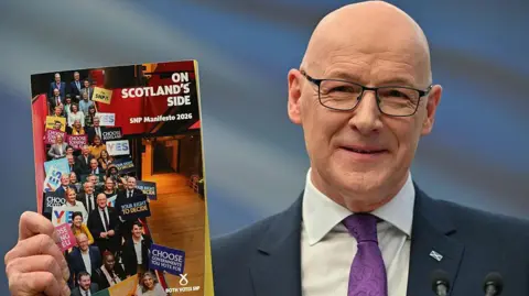 John Swinney, in dark blue suit with white shirt and purple tie, is holding up the SNP's election manifesto, which is called On Scotland's Side. He is smiling and wearing spectacles and a saltire badge on his left lapel. 