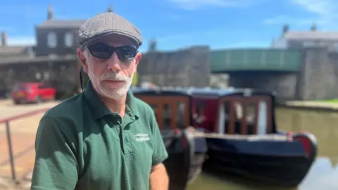 Olivia Richwald/BBC A man in a flat cap, sunglasses and green polo shirt stands in front of two out-of-focus narrow boats on a canal.