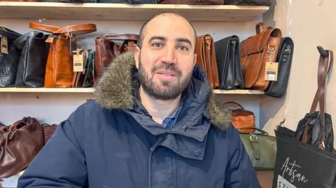 Noureddine is wearing a blue coat with fur collar and stood in front of a selection of handmade leather bags.