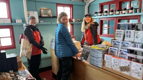 UK Antarctic Heritage Trust/ Lisa Ford A picture showing Dale Ellis with two female team mates. Dale is stood behind a desk in the shop with her team mates in front. There are postcards and cups on display. 