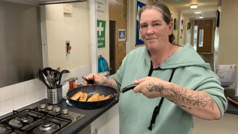BBC A woman is stood in a kitchen at a community centre. Her hair is tied back and she is wearing a green hoodie. She is holding up a frying pan full of sausages