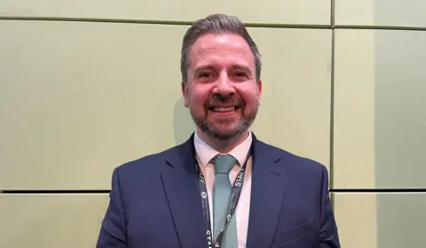 A man stands against a cream-coloured wall. He has grey hair and a grey beard. He is wearing a dark blue suit jacket, a pale pink shirt, and a pale green tie.