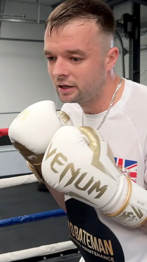 Jack Bateman stands in a boxing pose. He is wearing a white t-shirt with his name on and a union jack emblem. He is wearing white and gold boxing gloves on his hands. He has a silver chain around his neck. There are white and blue ropes of a boxing ring in the background.