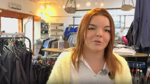 Marc Gaier/BBC A young woman with long light brown hair, wearig a white shirt and a yellow woolen cardigan, speaks to the camera in front of racks of school uniform clothing.