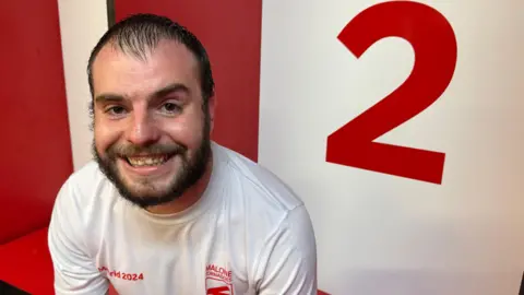 A man in a white t-shirt sitting down with a red number 2 behind him and red locker room wall. He has brown hair, with a brown beard and brown eyes. He is smiling at the camera. 
