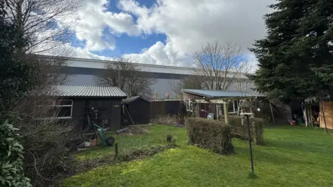 George King/BBC A large warehouse can be seem backing onto a back garden in which there are sheds, bird feeders, a wheelbarrow, and other gardening equipment.