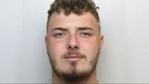 A police custody image of a man with short brown hair and a brown close-trimmed beard.