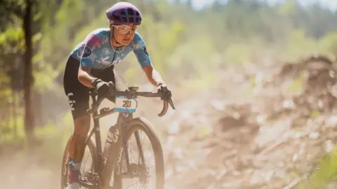 Red On Sports A gravel road racer in a blue and pink top and black shorts speeds through a dusty forest track