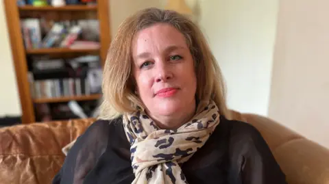 Kate Bradbrook/BBC A woman with long blonde hair, a leopard print scarf and a black top sits in a light brown chair in her home. 