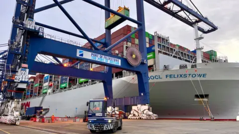 Martin Giles/BBC A small port vehicle is dwarfed by a crane that bears Port of Felixstowe branding. In the background there is a huge container ship, its deck packed with different coloured containers.