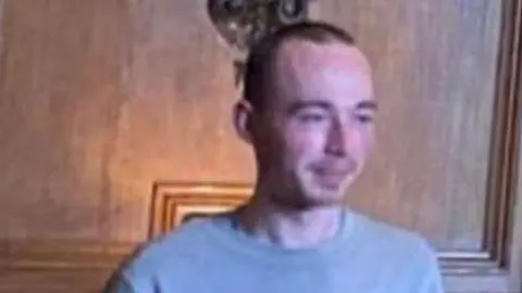 Family handout A man with short black hair wearing a blue jumper and standing in front of a wood panelled wall.