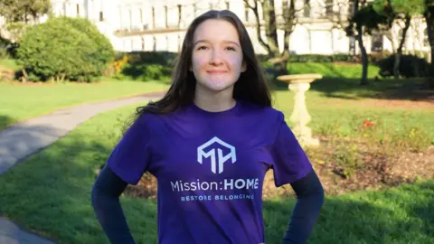 A teenage girl, wearing a dark purple t-shirt over a long sleeve black shirt. She is standing at a park.