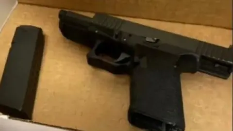 Obtained by CBS News Picture of a gun  that was found with Luigi Mangione. It is a pic of a black gun with a clip on the left of it. It appears to be on a piece of brown cardboard 