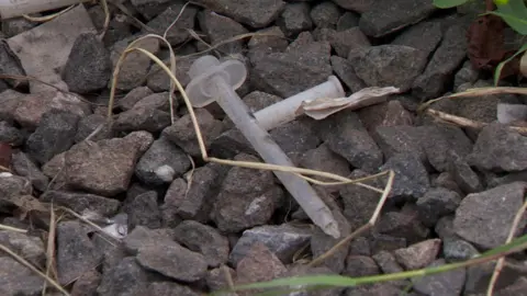 A used plastic needle lies discarded on the ground amongst stones