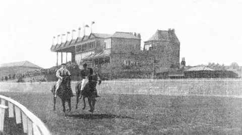 Grainy black-and-white photograph of two jockeys riding horses at New Barns in Salford. A large grandstand  with multiple flags on the roof, surrounded by a white picket fence, is in the background.
