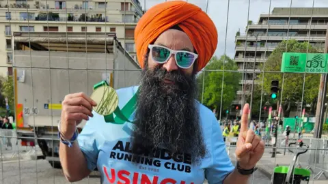 Kulwinder Cheema A man with a long black beard and moustache is wearing white sunglasses and an orange head turban. He is in a blight blur t-shirt and is holding a gold marathon medal with a green ribbon in one hand. The other hand is raised with one finger pointing up.
