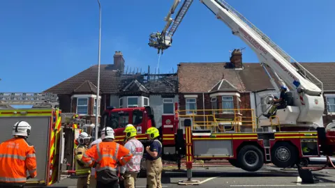 Five fightfighters in uniform with an aerial platform with a firefighter on it hosing down the fire, which is inside a terraced property with the roof burnt off.
There is a second fire engine in shot. The house is amongst a terraced row of houses. The sky is clear.
