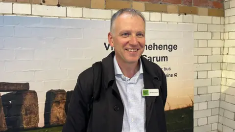 BBC Tim Hedley-Jones smiles while standing in front of subway tiles and a big picture of Stonehenge pointing to where the tour buses are