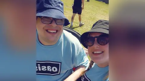 Jemma Tester Daniel Tester (left) wearing a sky blue Oasis t-shirt and navy blue bucket hat and sunglasses with his sister Jemma. She has blond plaits and is also wearing a sky blue Oasis t-shirt and black blue bucket hat and sunglasses at Heaton Park.