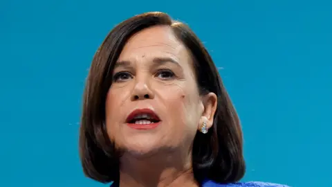 Reuters Sinn Fein President Mary Lou McDonald delivers a speech at the Sinn Fein annual party conference in Athlone. She has shoulder length brown hair and brown eyes.