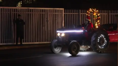 Joe Bilton / BBC A small tractor is leaving a compound. It has a man dressed as Santa and festive lights wrapped around a bar on the back of the vehicle. A green elf is also hanging from it.