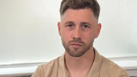 Billy Bashford has short blond hair and a goatee beard. He wears a camel-coloured polo shirt.