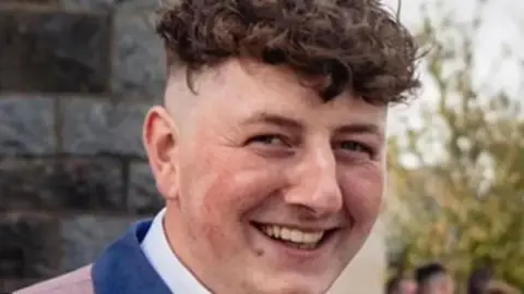 A man with short dark curly hair smiles towards the camera. It's closely cropped, but you can see the top of a pink jacket with navy blue collar and a white shirt. In the background is a building with grey stone and people in the distance. 