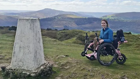 Bethany Handley Bethany in her all-terrain trike, which has pink metalwork, two large off-road wheels and a third motorised wheel at the back. She wears a blue down jacket and leggings with pink trainers. There is a trig point to the left of the trike. More hills can be seen in the background.