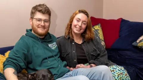 La Lotería del Código Postal del Pueblo James y su novia Rohanna sentados en un sofá azul. Ambos están sonriendo a la cámara. James tiene su mano sobre un perro que también está sentado en el sofá.