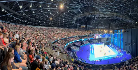 Ev Cargill A view from inside a sports arena, showing the thousands of people seated over three tiers to watch it. A netball court is on the ground, with teams in yellow and red playing each other.