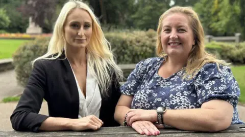 Two women seated at what appears to be a picnic table. The one on the left has long blonde hair and is wearing a black jacket and white blouse. The other has long fair hear and a blue and white short sleeved top