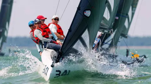 Cowes Week Paul Wyeth Crew members on a racing yacht lean over the side as the boat cuts through waves during a race.
