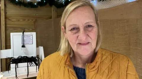Woman with short blonde hair wearing mustard yellow jacket with some cat diffuser products behind her and tinsel and Christmas lights above her head.