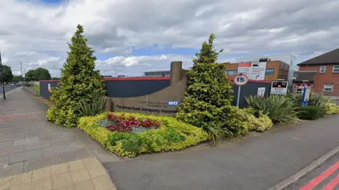 The entrance to the James Cook University Hospital. A wall displaying the hospital's name is surrounded by flowers and shrubs.