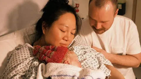 Serena Fan Serena and Benn are in a hospital room holding and looking down at a newborn Fenix who is wrapped in a blanket, with medical equipment and a wall clock visible behind them.