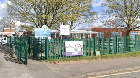 The exterior of a single storey school building fronted by green metal gates. There are two large trees in the school's front garden.  