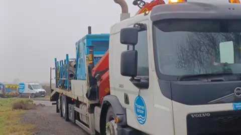 Thames Water A large Thames Water tanker truck with coiled pipework and big blue containers on the back. In the background there are more Thames Water vans and equipment. It is a grey and misty day.