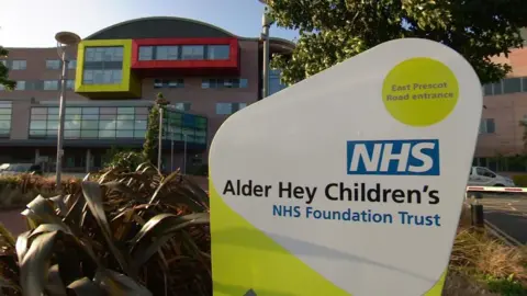 A sign outside the front of a large brick and glass building reads 'Alder Hey Children's NHS Foundation Trust'.