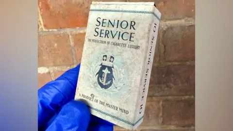 North Northamptonshire Council A Senior Service cigarette packet, mainly white with writing on it, begin held up by a person wearing blue gloves by a brick wall. 