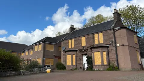 A large house with boarded-up windows underneath a bright blue sky