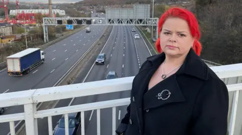 Ms Mercer stands on a bridge overlooking a smart motorway with traffic flowing in both directions. She has dyed red hair and is wearing a smart coat and necklace.