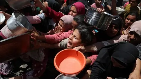 A large group of Palestinians wait to receive food cooked by a charity kitchen, in Jabalia, in the northern Gaza Strip.  They are holding on pans and other receptacles and look distressed. 