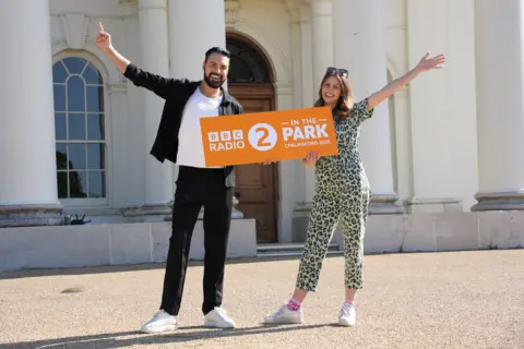 BBC Radio 2 Rylan Clark and Ellie Taylor stand in front of Hylands House, holding an orange sign with white writing which reads "BBC Radio 2 in the Park Chelmsford 2025". Rylan is stood on the left of frame, wearing black trousers and a black open shirt with white t-shirt underneath. He has dark hair and a beard. Ellie stands on the right of frame, wearing a leopard-print boiler suit, and has dark sunglasses pushed on top of her head. She has brown hair below shoulder-length. Both Rylan and Ellie are holding the sign with one hand and their other arm is spread wide. Hylands House behind them is a large white building with white columns.