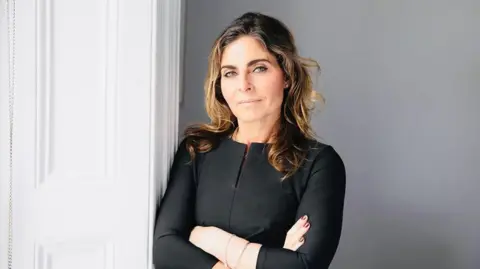 Laurie Fletcher A woman with long, wavy brown hair, wearing a smart black top. She is leaning against a white door with her arms folded and and is looking at the camera with a serious expression.