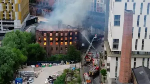 A drone aerial shot shows the fire raging inside the Hotspur building in Manchester city centre. An aerial ladder platform can be seen spraying water into the building, which sits in the shadow of large tower blocks and next to a number of trees.