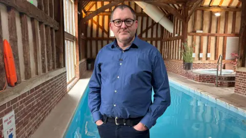 Daniel Pilley stands in front of the swimming pool in his health club, decorated with wooden beams and lights. The hot tub is in the background. He wears glasses, a blue shirt, and dark jeans.