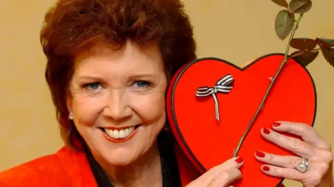 PA Cilla Black smiling and holding up a love heart while host of ITV show Blind Date